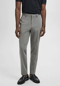 Graue Anzughose mit taillierter Passform, flachem Bund, Gürtelschlaufen und gebügelten Beinen. Getragen mit schwarzen Loafers und einem dunklen Oberteil.