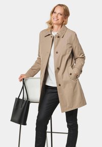 Beige dubbelzijdig jasje met een kraag, met knoopsluitingen en zijzakken. Gedragen over een witte top en zwarte strakke broek. Zwarte tote bag.