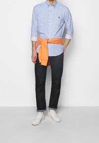 Jasnoniebieska koszula na guziki w kwiatowy wzór, z podwiniętymi rękawami, pomarańczowy sweter związany wokół talii, ciemne dżinsy i białe tenisówki.