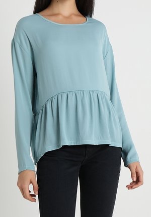 Kvinde iført en lyseblå bluse med lange ærmer og en samlet peplumkant samt sorte bukser mod en ensfarvet baggrund.