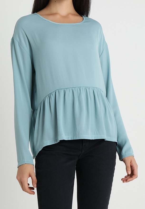 Femme portant un chemisier bleu clair à manches longues avec un ourlet péplum froncé et un pantalon noir sur un fond uni.