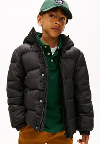 Chaqueta acolchada negra con capucha, cierre de cremallera, puños acanalados y bolsillos laterales. Llevada sobre una camisa polo verde. Tejido ligeramente texturizado.