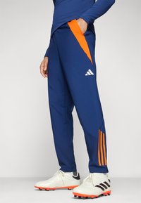 Pantaloni da atletica blu navy con accenti arancioni, dotati di tasche con zip e logo bianco. Indossati con scarpe chiodate bianche e arancioni.