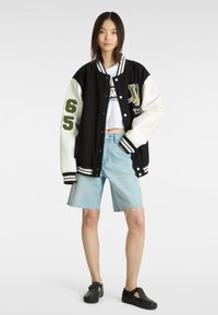 Chaqueta varsity en blanco y negro con bordado verde, camiseta blanca corta, pantalones cortos de mezclilla azul claro y zapatos negros con studs.