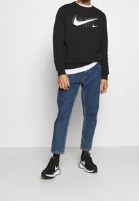 Musta paita valkoisella "AIR"-logolla, siniset farkut ja mustat Nike-tennarit. Yksinkertainen muotoilu, jossa resorit hihansuissa ja helmassa, klassinen löysä istuvuus.