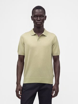Homme portant un polo beige clair à manches courtes et un pantalon foncé, debout devant un fond blanc uni.