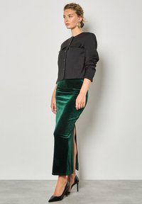 Giacca cropped nera con chiusura a bottoni e due tasche sul petto, abbinata a una gonna in velluto verde con spacco laterale. Scarpe con tacco nere.