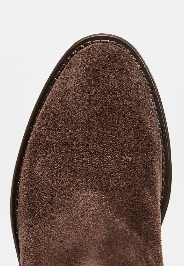 Brązowy zamszowy but z zaokrąglonym noskiem, kontrastującym przeszyciem wzdłuż podeszwy i gładką, teksturowaną powierzchnią.