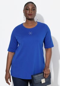 Zelfverzekerde vrouw in een koninklijk blauwe top met korte mouwen en kleine rhinestone details, grote oorbellen en een marineblauwe clutch met textuur in haar hand.