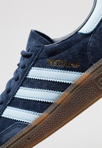 Tengerészkék velúr Adidas Spezial sneaker világoskék csíkkal, arany logóval és struktúrált áttetsző barna talppal, fehér háttér előtt, szögben állítva.