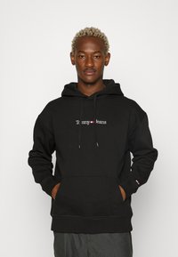 Felpa nera con cappuccio in misto cotone, dotata di tasca frontale a marsupio, cappuccio con stringhe, e logo bianco "Tommy Jeans". Polsini e orlo a costine.