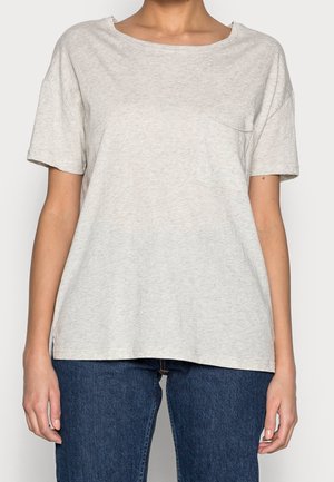 Vrouw die een lichtgrijs T-shirt met korte mouwen en een klein borstzakje draagt, gecombineerd met donkerblauwe spijkerbroek, staand tegen een gewone achtergrond.