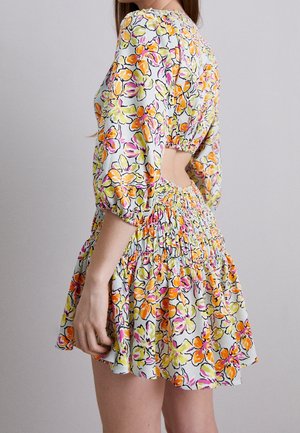 Kvinde iført en hvid kjole med pink, orange og gul blomsterprint, puffede ærmer og en udskæring på ryggen.