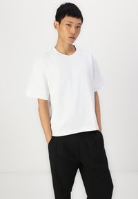 T-shirt bianca in cotone con maniche corte e scollatura rotonda, abbinata a pantaloni neri, su uno sfondo chiaro e uniforme.