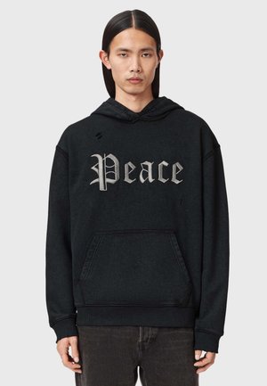 Ung person med langt, glat, mørkt hår iført sort hættetrøje med grå "Peace" tekst i gotisk skrift, kombineret med mørke bukser, neutral baggrund.