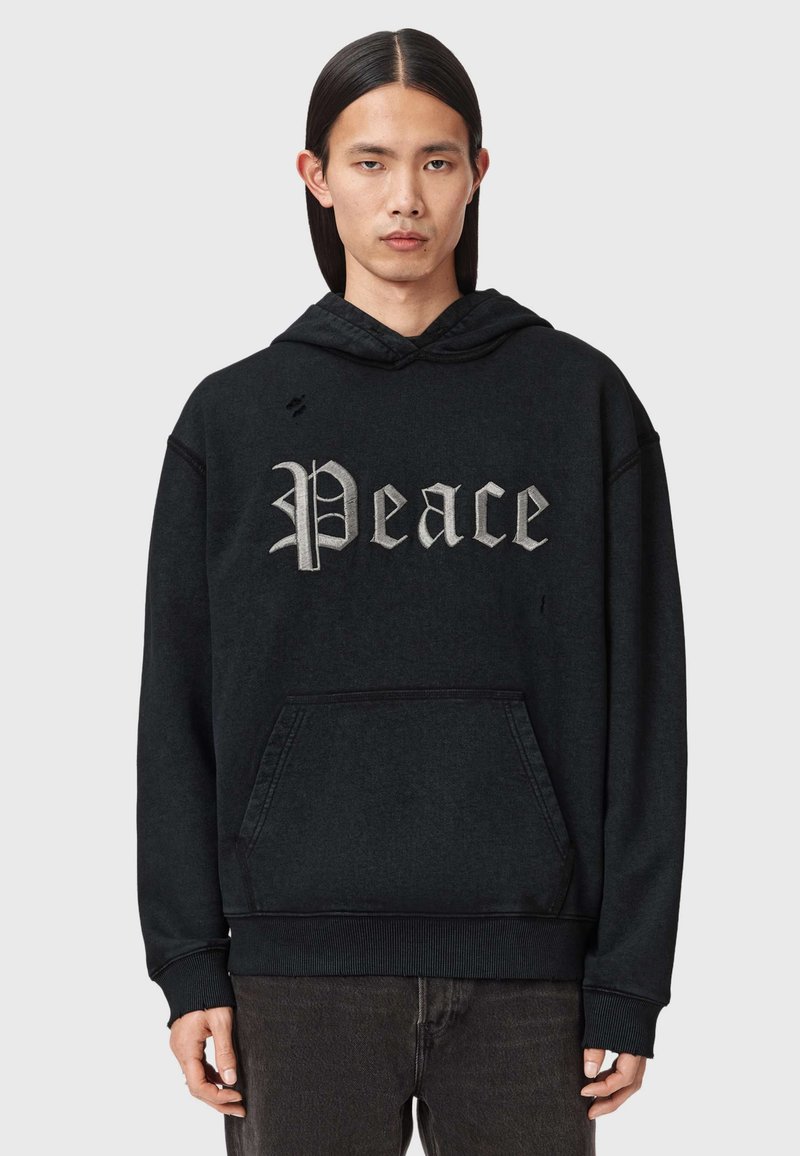 Jeune personne aux longs cheveux bruns lisses portant un sweat à capuche noir avec le texte gris "Peace" en police gothique, associé à un pantalon foncé, fond neutre.