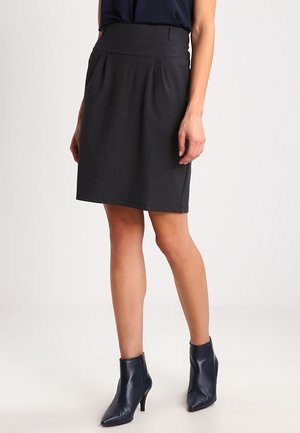 Femme portant une jupe gris foncé longueur genou, des bottines noires à talons hauts pointues et un haut foncé, debout devant un fond uni.