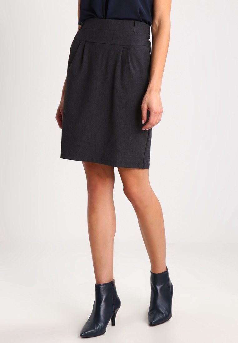 Femme portant une jupe gris foncé longueur genou, des bottines noires à talons hauts pointues et un haut foncé, debout devant un fond uni.