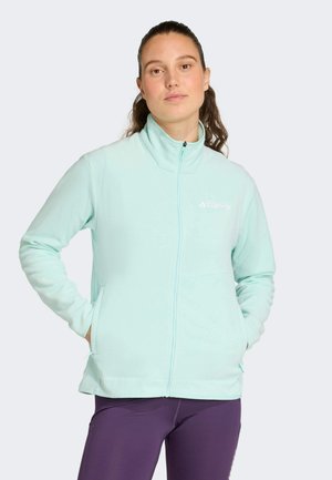 Frau trägt eine hellblaue Fleecejacke mit Reißverschluss und lila Leggings, steht mit den Händen in den Taschen vor einem schlichten Hintergrund.