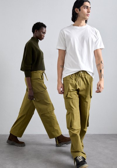Two models walk in olive green cargo pants, paired with a dark green sweater and a white t-shirt. Textured fabric with prominent pockets and drawstrings.