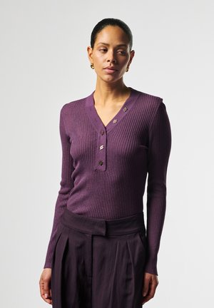 Vrouw met een paarse, geribbelde trui met lange mouwen en knopen en donkerpaarse, geplooide broek met hoge taille tegen een effen achtergrond.