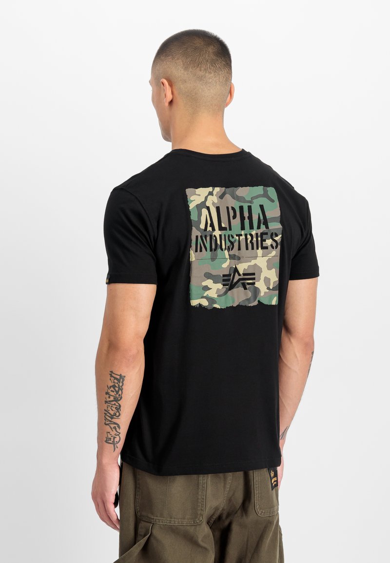 Άντρας με κοντά μαλλιά φοράει μαύρο t-shirt με το καμουφλάζ λογότυπο της Alpha Industries στην πλάτη, σε συνδυασμό με χακί cargos παντελόνια.