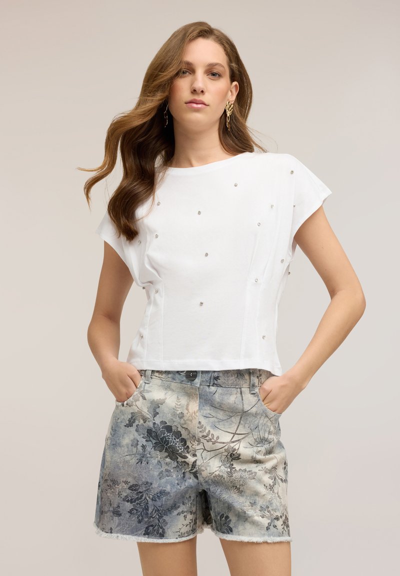 Young woman wearing a white short-sleeve top with small embellishments and floral-patterned denim shorts with hands in pockets.