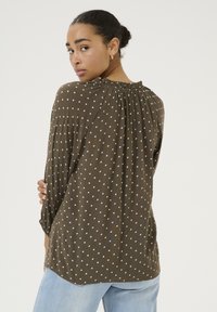 Blouse à pois marron avec un dos plissé, un col rond et des manches trois-quarts en tissu léger.