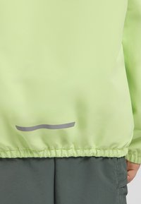 Giacca a vento di un verde chiaro con orlo arricciato e una striscia grigia come accentuazione. Tessuto liscio e polsini elasticizzati. Abbinata a pantaloni verdi scuri.