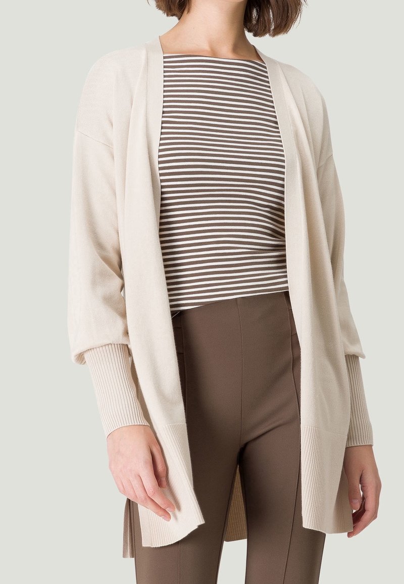 Femme portant un cardigan beige ouvert sur un haut rayé marron et blanc et un pantalon marron taille haute, sur un fond uni.