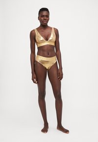 Zelta metāliska bikini komplekts, kas sastāv no V veida krūštura un augsto jostasvietu apakšbiksēm ar smalku dekoratīvu joslu. Gluda, atspulgojoša tekstūra.