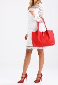 Punainen nahkainen käsilaukku, jossa on teksturoitu pinta ja suora siluetti. Varustettu kultasävyisellä metalliosalla ja yhdistetty valkoiseen pitsimekkoon.
