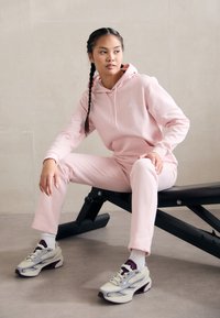 Hoodie rosa e calças de treino combinando, feitas de tecido suave, com um logótipo branco. Ténis bege e roxo com design texturizado. Sentado num banco.