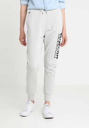 Osoba w jasnoszarych spodniach dresowych z napisem „NAUTI” na lewym udzie, białych sportowych butach i jasnoniebieskiej podwiniętej koszuli.