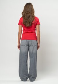 Pitkähiuksinen nainen, jolla on yllään punainen lyhythihainen paita ja väljät harmaat farkut, seisoo selin yksiväristä taustaa vasten.