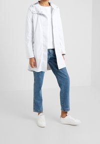 Cappotto impermeabile bianco con cappuccio, dotato di bottoni a pressione, abbinato a jeans blu a gamba dritta e sneakers bianche. Design semplice, texture liscia.