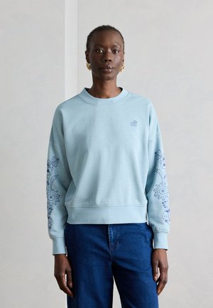 Femme portant un sweat-shirt bleu clair avec des motifs floraux bleus sur les manches et un jean bleu foncé, debout devant un fond gris uni.
