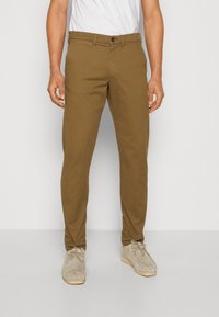 Pantaloni de bumbac maro cu o croială slim, având închidere cu buton și buzunare laterale. Asortați cu pantofi bej deschis pe un fundal simplu.
