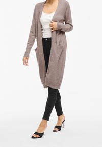 Kvinna klädd i en lång brun kofta, vit topp, svarta smala jeans och svarta öppen-tå hight heels som går på en vit bakgrund.