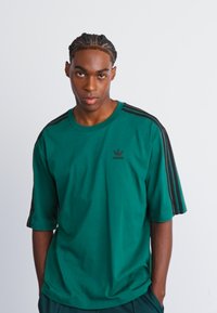 Grøn kortærmet t-shirt lavet af bomuld med sorte striber på skuldrene og et lille sort Adidas-logo på brystet. Glat tekstur.