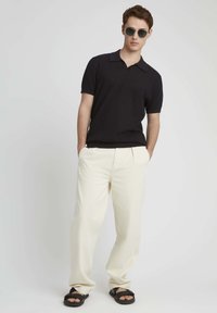 Polo shirt noir à manches courtes avec un tissu texturé, associé à un pantalon crème ample et des sandales noires à enfiler.