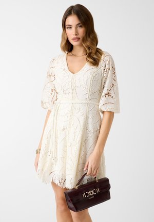 Kobieta ubrana w białą koronkową sukienkę z bufkami, trzymająca ciemnobrązową torebkę Guess z uchwytem od góry na jednolitym tle.