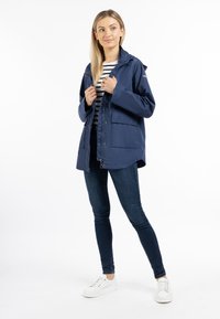 Giacca impermeabile blu navy con cappuccio, cerniera frontale, due grandi tasche frontali e un design leggermente oversize. Indossata sopra una maglietta a righe e un paio di jeans.