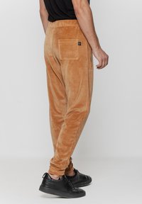 Beżowe welurowe dresy o luźnym kroju, z boczną kieszenią na zamek i tylną kieszenią naszywaną, w połączeniu z czarnymi sneakersami, prezentujące gładką teksturę.