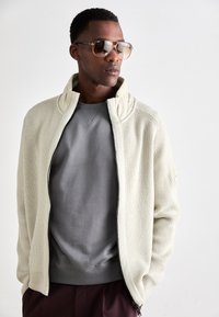 Veste en tricot zippée crème avec col haut, associée à un sweat-shirt gris. Le modèle porte des lunettes de soleil avec des accents dorés. Pantalons bordeaux foncé.