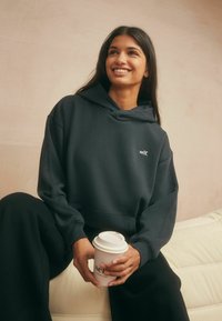 Donkergrijze hoodiesweater met het "self." logo, gemaakt van zachte stof. Het model houdt een koffiekop vast terwijl hij/zij op een gestructureerd oppervlak zit.