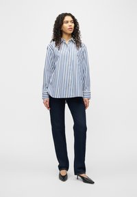 Γυναίκα που φοράει μπλε και λευκό πουκάμισο με κάθετες ρίγες, σκούρο μπλε τζιν και μαύρες μυτερές γόβες, στέκεται σε λευκό φόντο.