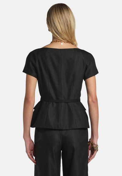 Frau mit blondem Haar, die ein eng anliegendes schwarzes Kurzarmoberteil und passende schwarze Hosen trägt, von hinten vor weißem Hintergrund gesehen.