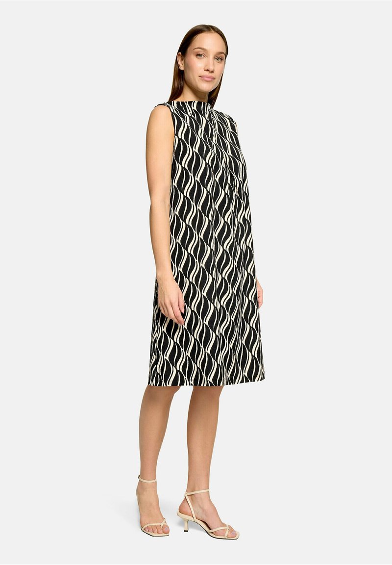 Femme debout portant une robe sans manches noire et blanche à motif ondulé, longueur genou, et des talons beiges à brides sur un fond uni.
