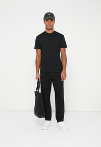 Hombre de pie contra un fondo blanco, llevando una gorra negra, una camiseta negra, pantalones negros, zapatillas blancas y sosteniendo una bolsa tote negra.
